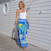 blonde woman in white one shoulder swimsuit with blue palm print wrap skirt coverup